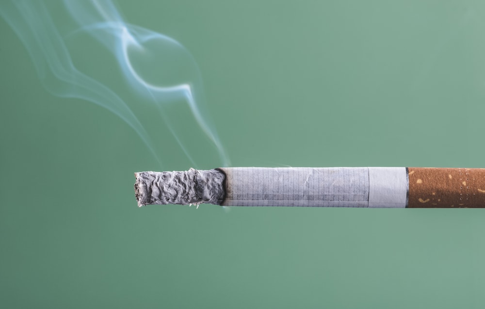 Cigarette burning with smoke on green background close up, quit smoking concept